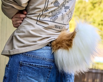 May include: A fluffy, faux fur tail in white and brown, attached to the back of blue jeans. The tail has a brown patch at the base and transitions to white. The person is wearing a tan sweatshirt with the word "MIRACLE" printed on the back.