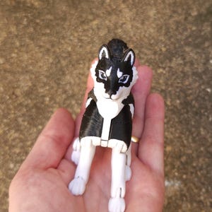Puede incluir: Una figura de husky en blanco y negro con articulaciones. El perro de juguete tiene la cabeza y la espalda negras con detalles blancos en la cara, el pecho y las patas. La figura se sostiene en una mano.