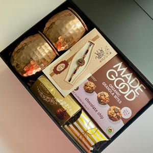 May include: A black gift box filled with two hammered copper mugs, a box of chocolate chip granola bites, a Lindor chocolate bar, and a decorative bracelet. The box also contains a gold and white striped package.