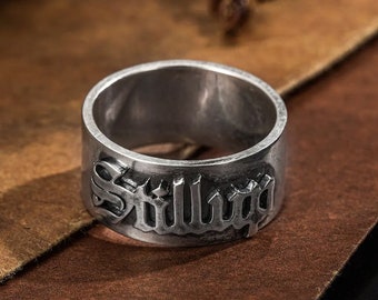 May include: A silver-colored ring with the word "Stillboy" in gothic lettering. The ring has a wide band and a slightly worn appearance. The ring is displayed on a brown surface.