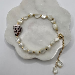 May include: A delicate bracelet featuring white, irregularly shaped beads and small gold-colored accents. A conch shell with brown and black spots is a focal point. The bracelet has a gold chain with two dangling shell-shaped charms.