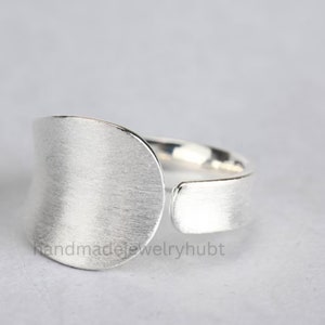 May include: A silver ring with a wide, flat band and a curved, oval-shaped top. The ring has a brushed finish.