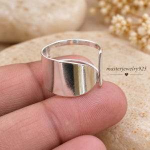 Könnte beinhalten: Ein polierter Silberring mit einem breiten, offenen Banddesign und einer glatten, reflektierenden Oberfläche. Der Ring zeigt eine geschwungene, überlappende Struktur und enthält den Text masterjewelry925 auf der rechten Seite des Bildes.