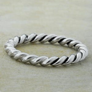 Könnte beinhalten: Ein silberfarbener Ring mit einem gedrehten Seildesign. Der Ring besteht aus glänzendem Metall und hat eine durchgehende, kreisförmige Form. Das gedrehte Muster erzeugt eine strukturierte Oberfläche.