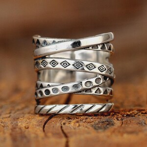 May include: A silver-colored ring with an intricate design. The ring features multiple bands that wrap around each other, adorned with geometric patterns like triangles, circles, and diamonds. The ring is set against a blurred wooden background.