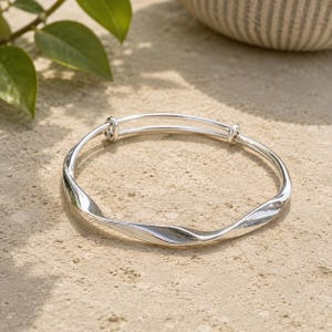 Può includere: Un bracciale rigido estensibile color argento con un design in metallo ritorto e ondulato. Il bracciale presenta due estremità sovrapposte che scorrono attraverso due piccoli anelli metallici, consentendo di regolare il diametro per il polso.