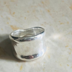 May include: A silver ring with a thick, rounded band and a slightly uneven surface. The ring reflects light and has a simple, modern design. The ring's shape is not perfectly circular. It is likely made of silver.