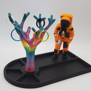 May include: A 3D printed astronaut figurine stands next to a rainbow-colored tree with multiple rings hanging from its branches. The astronaut and tree are on a black tray with multiple compartments.