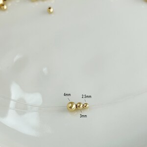 May include: A close-up of a gold-colored necklace with three beads of varying sizes: 4mm, 3mm, and 2.5mm. The necklace is on a clear string, and the beads are scattered on a white surface.