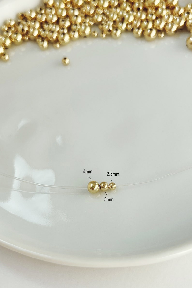 May include: A close-up of a gold-colored necklace with three beads of varying sizes: 4mm, 3mm, and 2.5mm. The necklace is on a clear string, and the beads are scattered on a white surface.