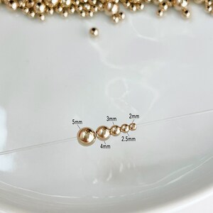 May include: Gold-colored beads of varying sizes, from 2mm to 5mm, are strung on a clear thread. The beads are displayed on a white surface, with a scattering of similar beads in the background. The image includes size labels for each bead.