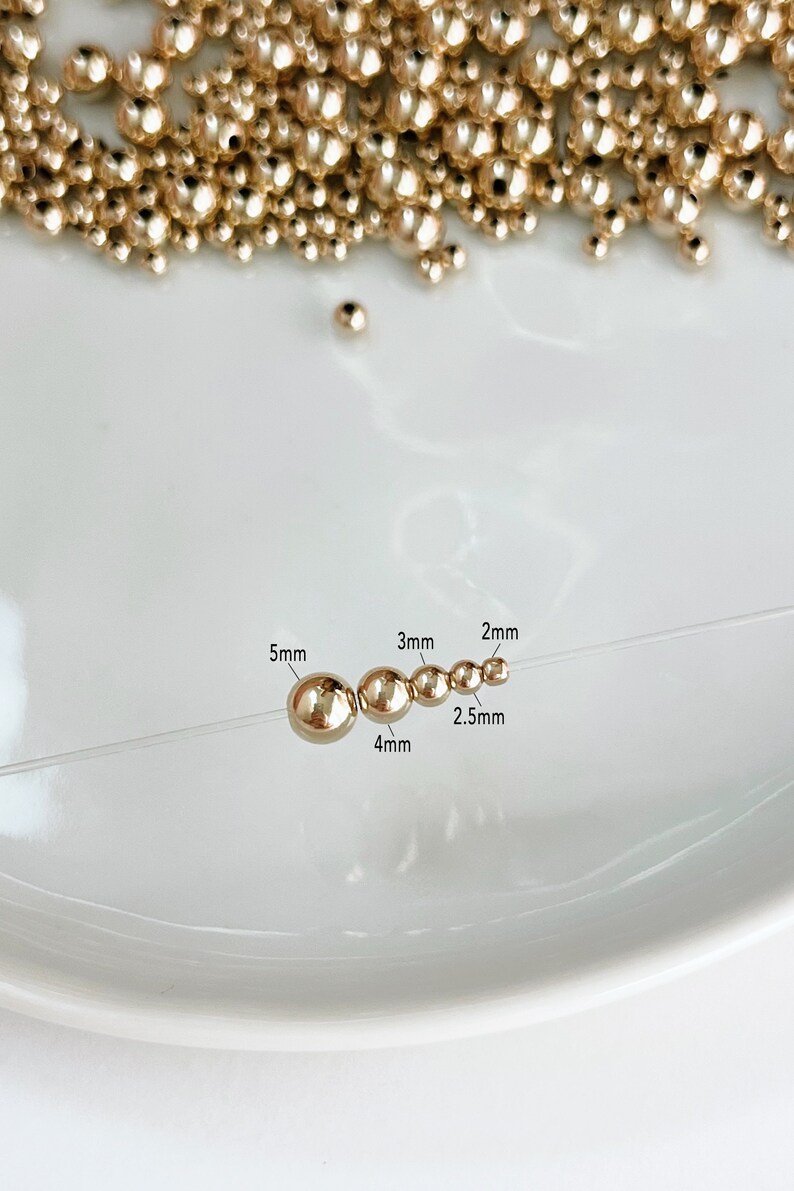 May include: Gold-colored beads of varying sizes, from 2mm to 5mm, are strung on a clear thread. The beads are displayed on a white surface, with a scattering of similar beads in the background. The image includes size labels for each bead.
