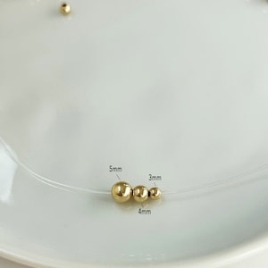 May include: A delicate necklace featuring three gold-colored beads, sized 5mm, 4mm, and 3mm, strung on a clear cord. The beads are displayed on a white plate, with additional gold beads scattered in the background. The necklace is a minimalist jewelry piece.