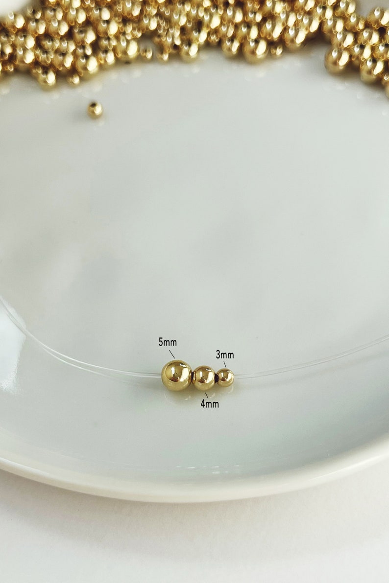 May include: A delicate necklace featuring three gold-colored beads, sized 5mm, 4mm, and 3mm, strung on a clear cord. The beads are displayed on a white plate, with additional gold beads scattered in the background. The necklace is a minimalist jewelry piece.