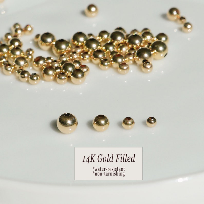 May include: A collection of 14K gold-filled beads in various sizes, scattered on a white surface. The beads are round and shiny, reflecting light. The image includes text that reads "14K Gold Filled", "water-resistant", and "non-tarnishing".