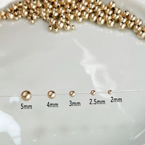 May include: Gold-colored round beads in various sizes, ranging from 2mm to 5mm, displayed on a white surface. The sizes are labeled: 5mm, 4mm, 3mm, 2.5mm, and 2mm. A collection of similar beads is scattered in the background.