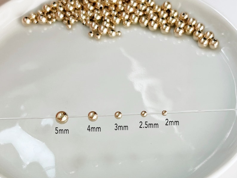 May include: Gold-colored round beads in various sizes, ranging from 2mm to 5mm, displayed on a white surface. The sizes are labeled: 5mm, 4mm, 3mm, 2.5mm, and 2mm. A collection of similar beads is scattered in the background.