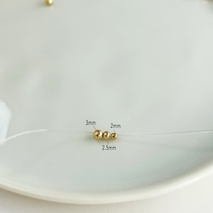May include: Close-up of gold-colored beads on a clear string, with three beads in focus. The beads are 3mm, 2mm, and 2.5mm in diameter. The beads are on a white plate, with more gold beads out of focus in the background.