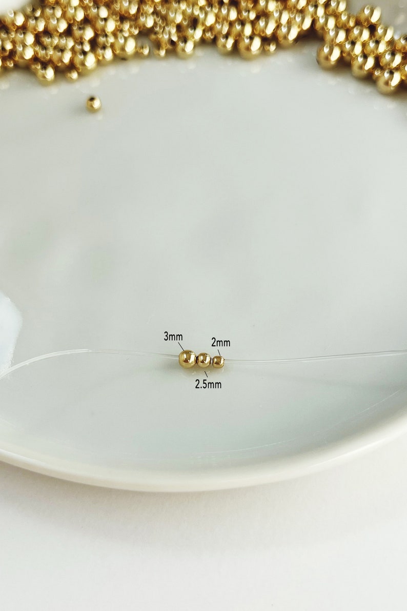 May include: Close-up of gold-colored beads on a clear string, with three beads in focus. The beads are 3mm, 2mm, and 2.5mm in diameter. The beads are on a white plate, with more gold beads out of focus in the background.