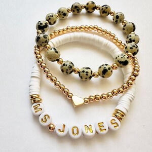 May include: A set of three stacked bracelets. One features dalmatian-print stone beads. Another is a strand of small gold-tone metallic beads with a central gold heart charm. The third is white disc beads with gold-tone letter beads spelling MS JONES.