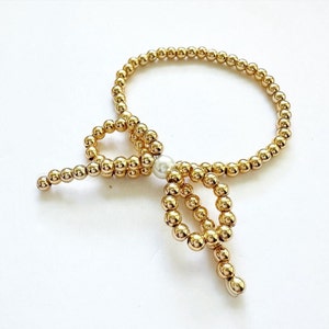 May include: A gold beaded bracelet with a pearl accent and a bow-shaped design.