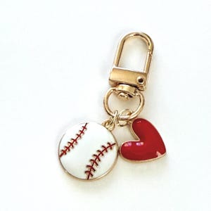 May include: Gold-tone keychain with a baseball charm and a red heart charm. The baseball charm is white with red stitching. The heart charm is a glossy red. The keychain has a lobster clasp.