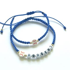 May include: Two blue braided bracelets. One bracelet features a baseball charm and the other spells out "DODGERS" in white letter beads. The bracelets have adjustable blue cords and gold-colored accents.