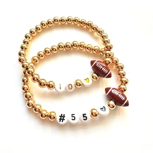 May include: Two gold beaded bracelets. One bracelet spells "I 0" with a yellow heart bead. The other bracelet spells "# 5 5" with a blue heart bead. Each bracelet has a brown football charm.