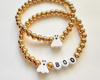 May include: Two gold beaded bracelets with white ghost charms and the word "BOO" spelled out in black and white letter beads. The bracelets are on a white surface.