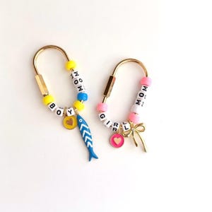 May include: Two gold keychains with beaded accents. One keychain says "BOY" and features a blue fish charm and a yellow heart charm. The other says "GIRL" and has a gold bow and pink heart charm.