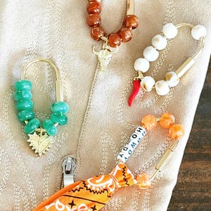 May include: Four decorative keychains with gold-colored hardware. One keychain has green beads and a gold heart charm. Another has brown beads and a bull skull charm. A third has white beads and a red chili pepper. The last has orange beads and an orange bandana with the word "HOWDY".