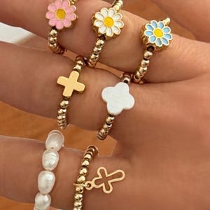 May include: A collection of beaded rings in gold tones, featuring various designs. Some rings have flower-shaped embellishments in pink, white, and blue. Other rings showcase a gold cross, a white clover, and a pearl band. The rings are displayed on a hand.