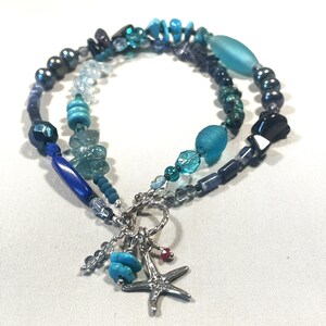 Op de afbeelding: Een gelaagde armband met blauwe en turquoise kralen in verschillende vormen. Een zilveren zeesterbedel en turquoise kralen hangen aan de armband, wat een nautisch thema creëert. De armband is ongeveer 19 cm lang.