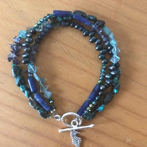 Op de afbeelding: Een armband met meerdere strengen met blauwe en turquoise edelstenen, waaronder turkoois, lapis lazuli en facetkralen. De armband heeft een zilveren tuimelsluiting met een zeepaardjesbedel. De armband wordt gepresenteerd op een houten oppervlak.