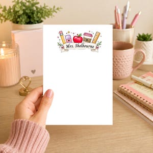 Op de afbeelding: Een wit notitieblok met een kleurrijke illustratie van schoolbenodigdheden. Het ontwerp bevat een potlood, appel, koffiekopje en liniaal, met de tekst "Mrs. Shelbourne" op een banner.