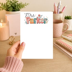 Puede incluir: Un bloc de notas blanco con el texto "Mrs. Parker" en letras coloridas y decorativas. El bloc de notas está sostenido por una persona, con una vela, una taza y artículos de papelería en el fondo.