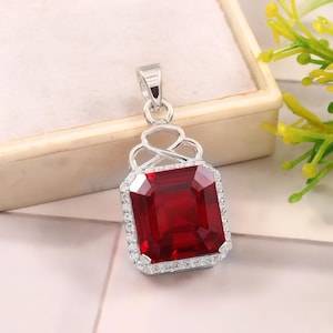 May include: A silver-toned pendant featuring a large, rectangular, deep red gemstone set in a frame of small, clear, sparkling stones. The pendant has an intricate, open-work silver bail at the top, connecting to a polished silver loop for a chain.