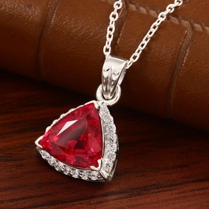 Può includere: Una collana d'argento con un ciondolo triangolare con una gemma rossa brillante. Il ciondolo è circondato da un alone di piccoli cristalli trasparenti. La catena è sottile e argentata, creando un elegante gioiello.