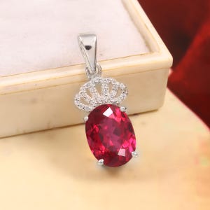 May include: A silver pendant featuring a large, oval-cut, deep red gemstone set in a prong setting. Above the stone is a decorative silver crown-shaped bail encrusted with small, clear, sparkling stones, attached to a polished silver loop.