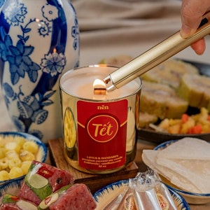 May include: A gold candle with a red label that says "nên Tết" and "LOTUS & MANDARIN premium coconut soy candle California | ameliehome.com 10 oz" is being lit with a gold lighter. The candle is surrounded by various snacks, including a bowl of yellow and orange snacks, a bowl of red and green snacks, and a bowl of clear wrapped snacks.