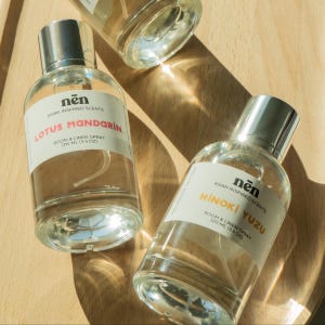 May include: Three clear glass spray bottles with silver caps are arranged on a wooden surface. Each bottle has a white label with the brand name "nën" and the words "Asian Inspired Scents." The labels read "Lotus Mandarin" and "Hinoki Yuzu" with "Room & Linen Spray 100 ml (3.4 oz)."
