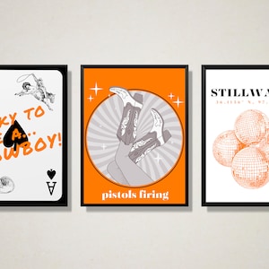 May include: Three framed prints with orange backgrounds and black frames. The left print features a black ace of spades playing card with the text "Lucky to be a... Cowboy!" The middle print features a pair of cowboy boots with the text "pistols firing". The right print features the text "Stillwater 36.1156 N. 97.0584 W" with a cluster of disco balls.