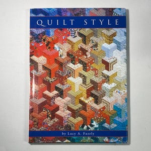 Peut inclure: Un livre rectangulaire intitulé Quilt Style par Lucy A. Fazely. La couverture présente un motif géométrique multicolore et vibrant de cubes empilés dans des tons de rouge, orange, jaune, marron et bleu, créant une illusion d'optique en 3D.