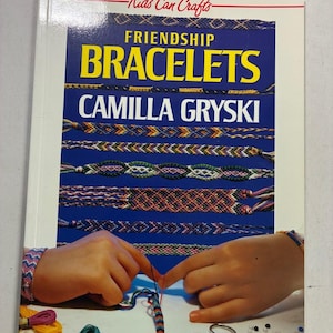 May include: A softcover instructional book titled Kids Can Crafts Friendship Bracelets by Camilla Gryski. The cover features a blue background with various woven thread bracelets, showing hands creating a bracelet with colorful embroidery floss.