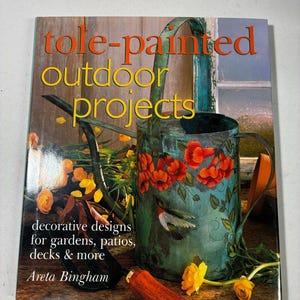 May include: A softcover book titled tole-painted outdoor projects by Areta Bingham. The cover features a teal metal watering can decorated with orange trumpet vine flowers and a hummingbird, alongside a garden trowel and yellow flowers.