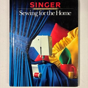 Peut inclure: Un livre de la bibliothèque de référence de couture Singer intitulé Sewing for the Home. La couverture présente une machine à coudre blanche, des rideaux bleus, un coussin rouge, un coussin jaune à volants, une serviette verte dans une tasse rouge.