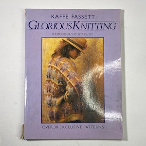 May include: A softcover book titled Glorious Knitting by Kaffe Fassett with photography by Steve Lovi. The lavender cover features a photograph of a person wearing a multi-coloured patterned jumper and a straw hat, with the text Over 30 Exclusive Patterns.