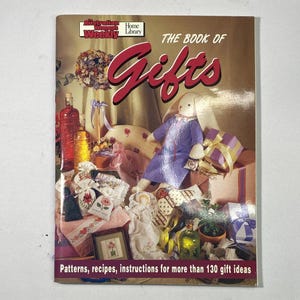 May include: A softcover book titled The Australian Women's Weekly Home Library The Book of Gifts. The cover features a stuffed rabbit in a purple dress, various gift-wrapped boxes, glass bottles, and embroidered fabric items on a table.