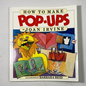 May include: A softcover book titled How to Make Pop-Ups by Joan Irvine, illustrated by Barbara Reid. The cover features a yellow lion, a ghost behind a door, and a person wearing a red crown, all rendered in a colorful, hand-drawn art style.