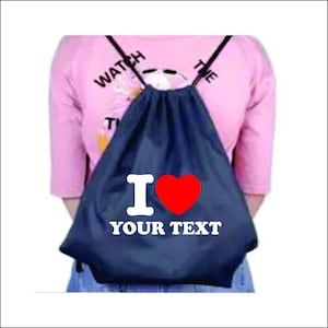 May include: A dark blue drawstring backpack featuring a white letter I, a red heart symbol, and the white text YOUR TEXT printed on the front. The bag has black cord straps and is worn over a light pink shirt with partial text visible at the top.